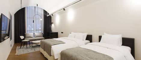 A modern hotel room with two beds, a sofa, a desk, and a city view.