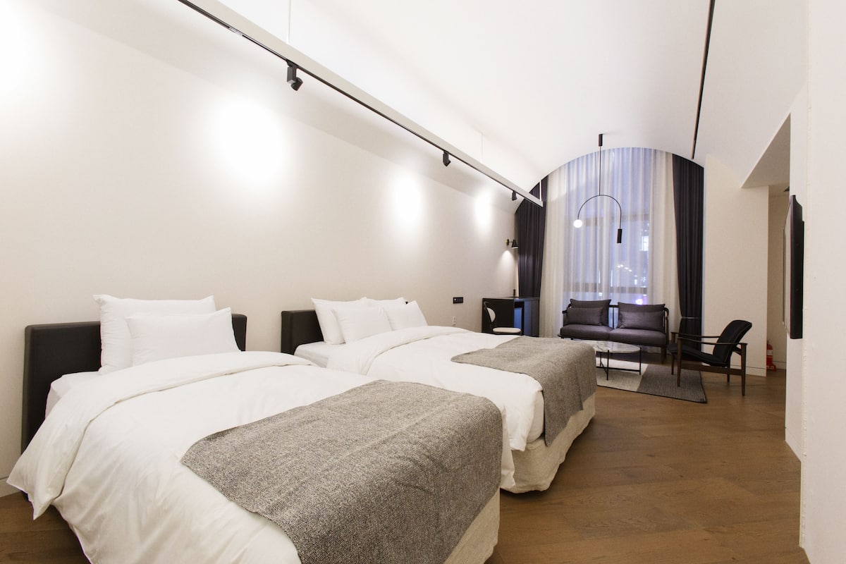 Habitación de hotel moderna con dos camas, un sofá, una mesita y vistas a la ciudad a través de un ventanal.