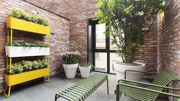 A brick-walled courtyard with a yellow planter, two white planters, a bench, and two green chairs.