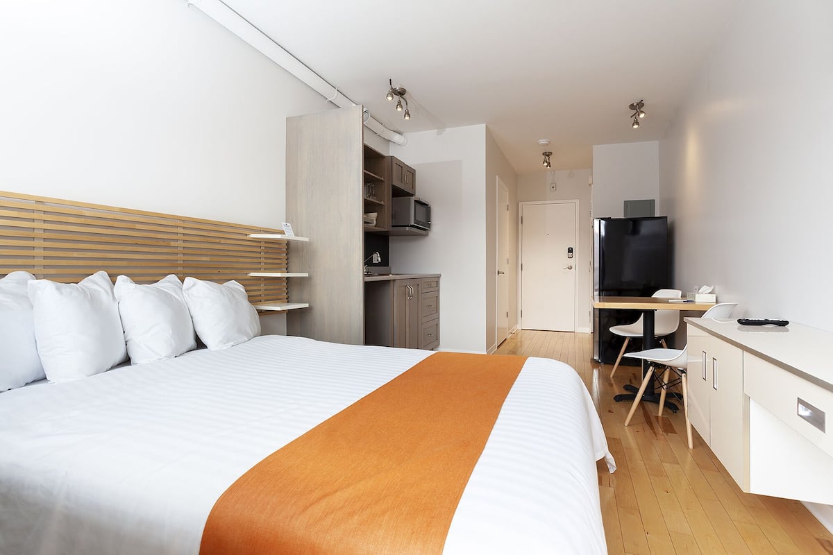 A hotel room with a bed, a desk, a chair, a TV, and a kitchenette.