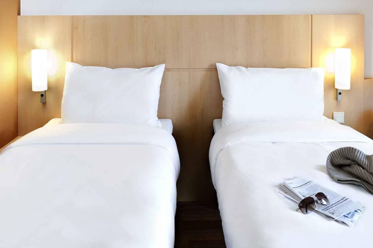 A hotel room with two single beds, each with a white mattress and pillow, and a wooden headboard. There are wall-mounted light fixtures on either side of the headboard.