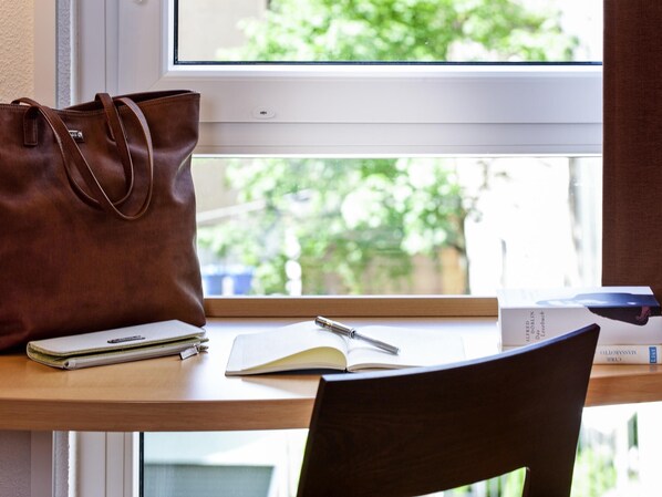 Ein Schreibtisch mit einer braunen Ledertasche, einem Notizbuch, einem Stift und einem Buch mit dem Titel 'VIA D'ALBERTO' auf einer Holzoberfläche in der Nähe eines Fensters.