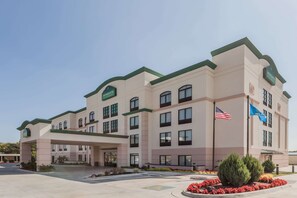 Un hotel de varios pisos con fachada verde y beige, una entrada cubierta y las banderas del estado de Oklahoma y de Estados Unidos.