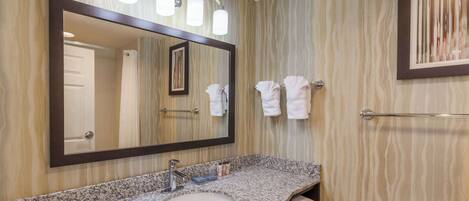 A bathroom with a granite countertop sink, a large mirror with lights, a towel rack, and a framed picture on the wall.
