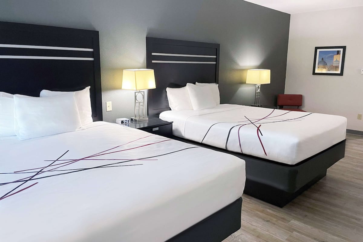 A hotel room with two beds, each with a black headboard and white bedding. There are bedside tables with lamps, a red chair, and a framed picture on the wall.