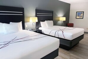 Habitación de hotel con dos camas, cada una con cabecero negro y ropa de cama blanca. Se observan mesitas de noche con lámparas, una silla roja y un cuadro enmarcado en la pared.