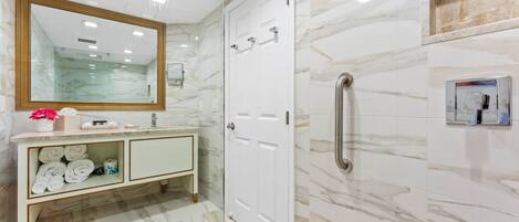A bathroom with a marble floor, a vanity with a mirror, and a shower area with a grab bar.