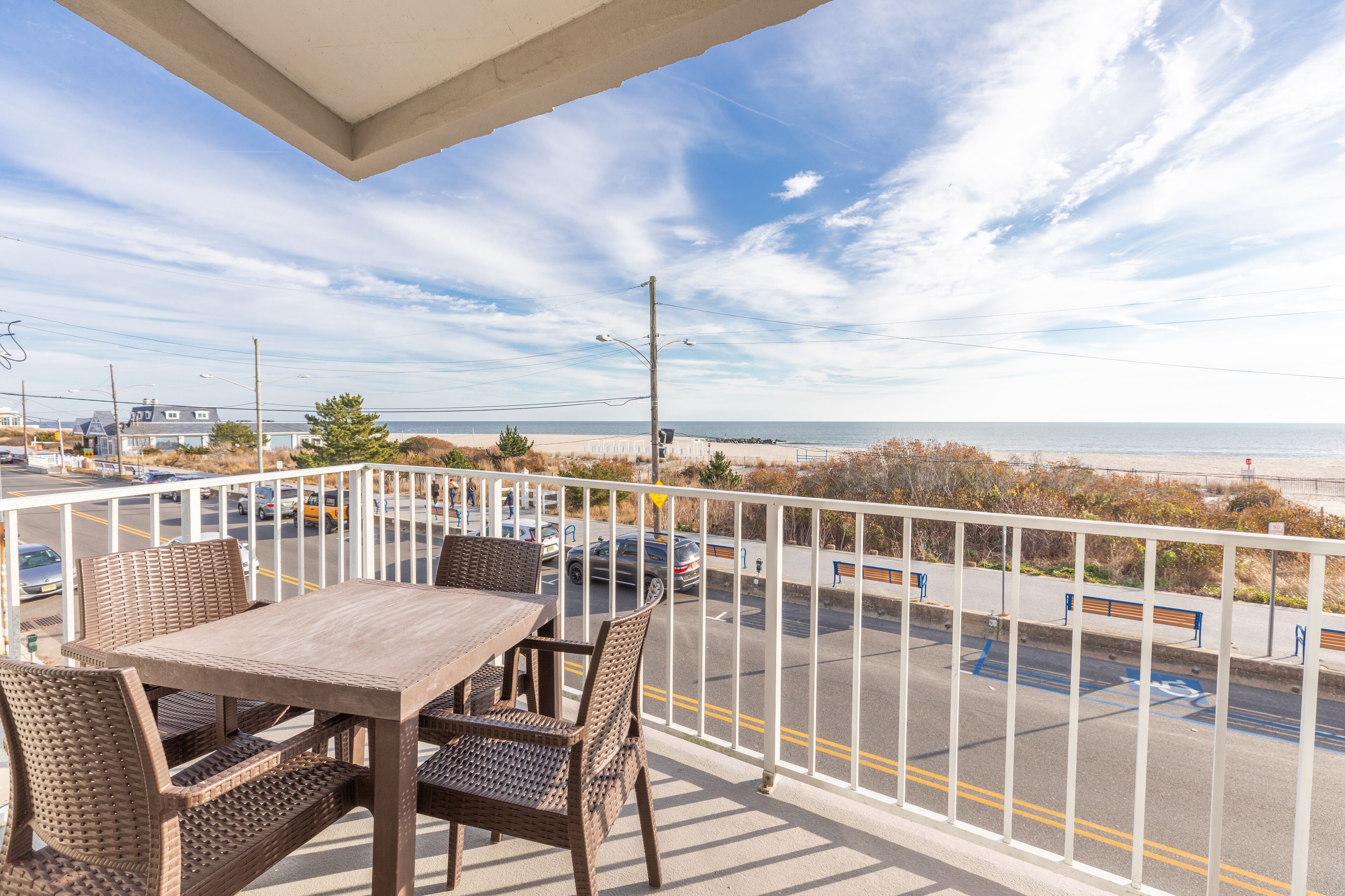A balcony with a table and chairs overlooking a parking lot and beach.