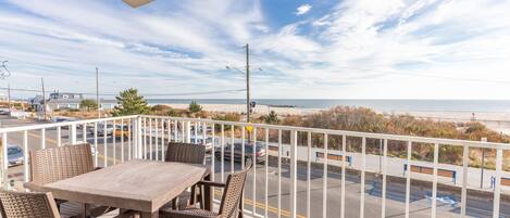 A balcony with a table and chairs overlooking a parking lot and beach.
