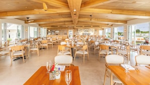 Un restaurante espacioso con altos techos de madera y ventanas amplias que ofrecen vistas al exterior. Las mesas están dispuestas con sillas blancas y copas de vino.