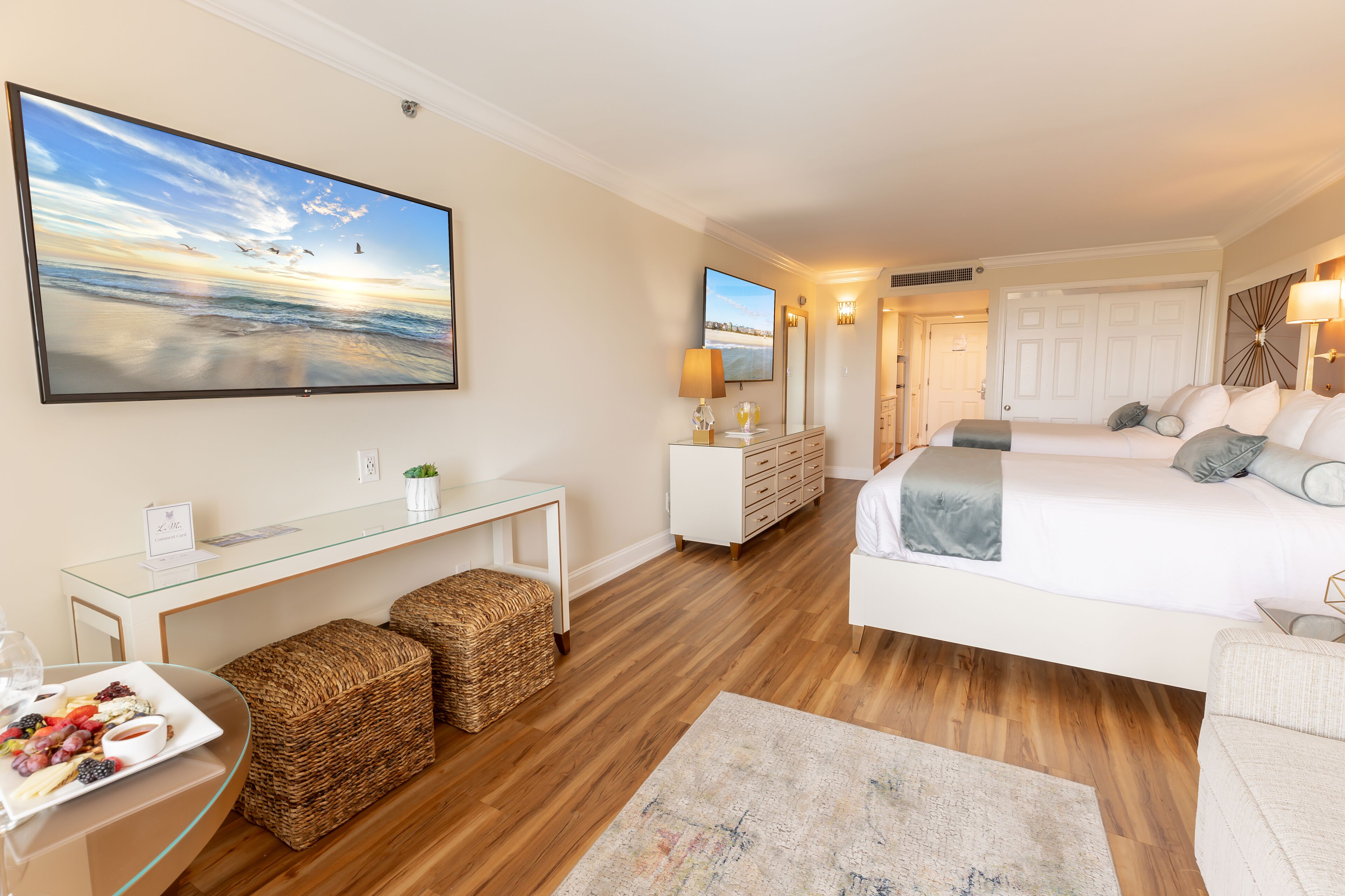 A modern hotel room with a large flat-screen TV, a bed, a sofa, a glass coffee table with fruit, and wicker baskets.