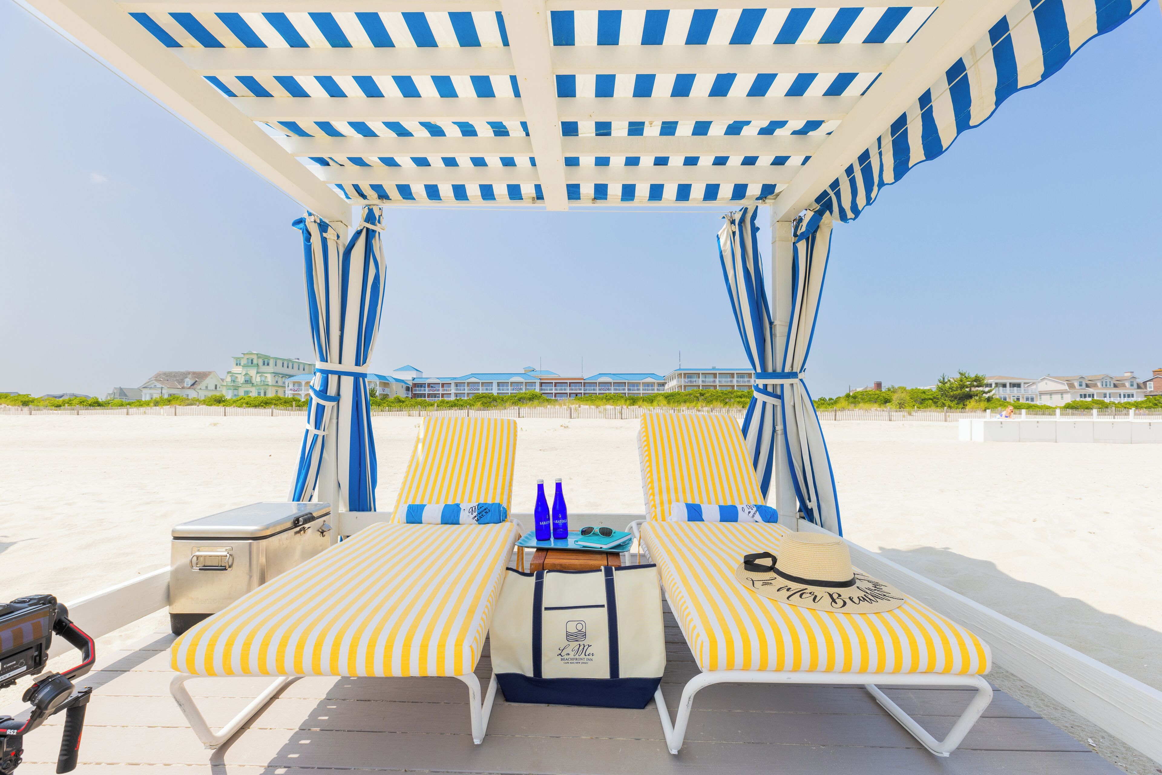 Un espacio de descanso frente al mar con sillones reclinables a rayas amarillas y blancas, una mesa con botellas y gafas de sol, y un toldo por encima.