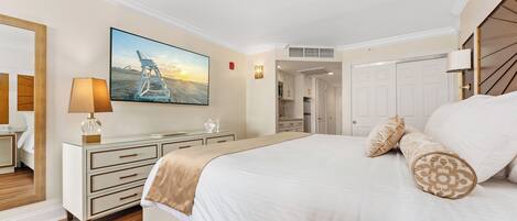 Une chambre d’hôtel avec un grand lit, une table de chevet avec une lampe, un tableau représentant une scène de plage, et une vue sur la cuisine et la salle de bain par une porte ouverte.