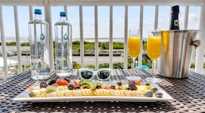 A breakfast setup with a plate of food, two bottles of water, two glasses of orange juice, and a bottle of champagne on a balcony with a view.