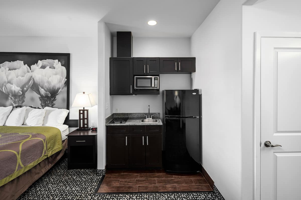 A compact hotel room with a bed, a small kitchenette, a microwave, a refrigerator, and a nightstand with a lamp.