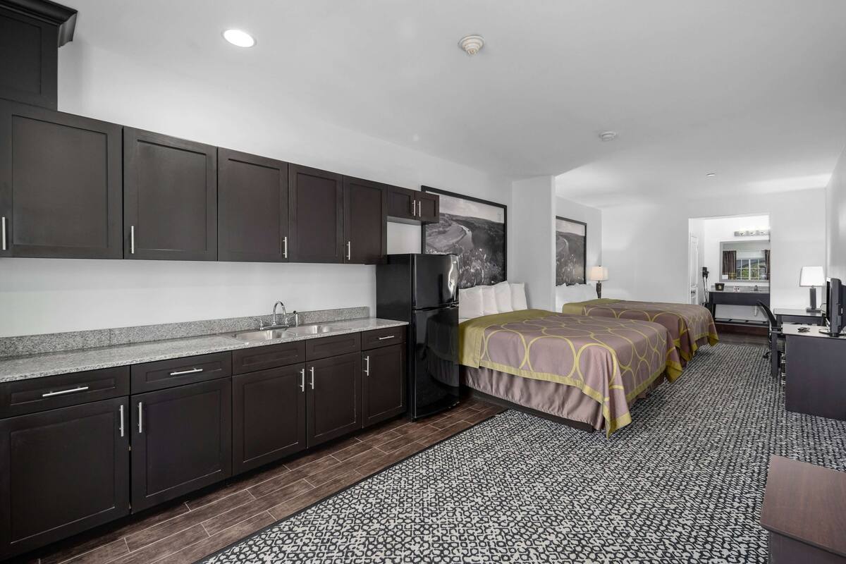 A hotel room with a kitchenette, two beds, a desk, and a bathroom.