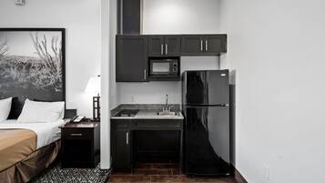 A compact hotel room with a kitchenette, a bed with a headboard, a nightstand, and a wall-mounted picture.