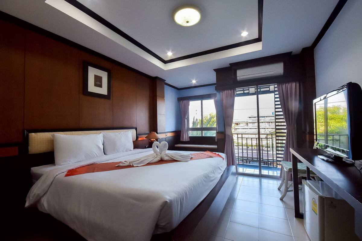 A hotel room with a large bed, a desk with a computer, a window with curtains, and a balcony with a view.
