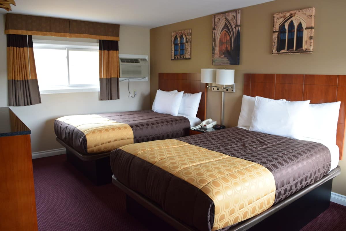 A hotel room with two beds, a wooden headboard, a window with curtains, a wall-mounted air conditioner, and two wall-mounted lamps.