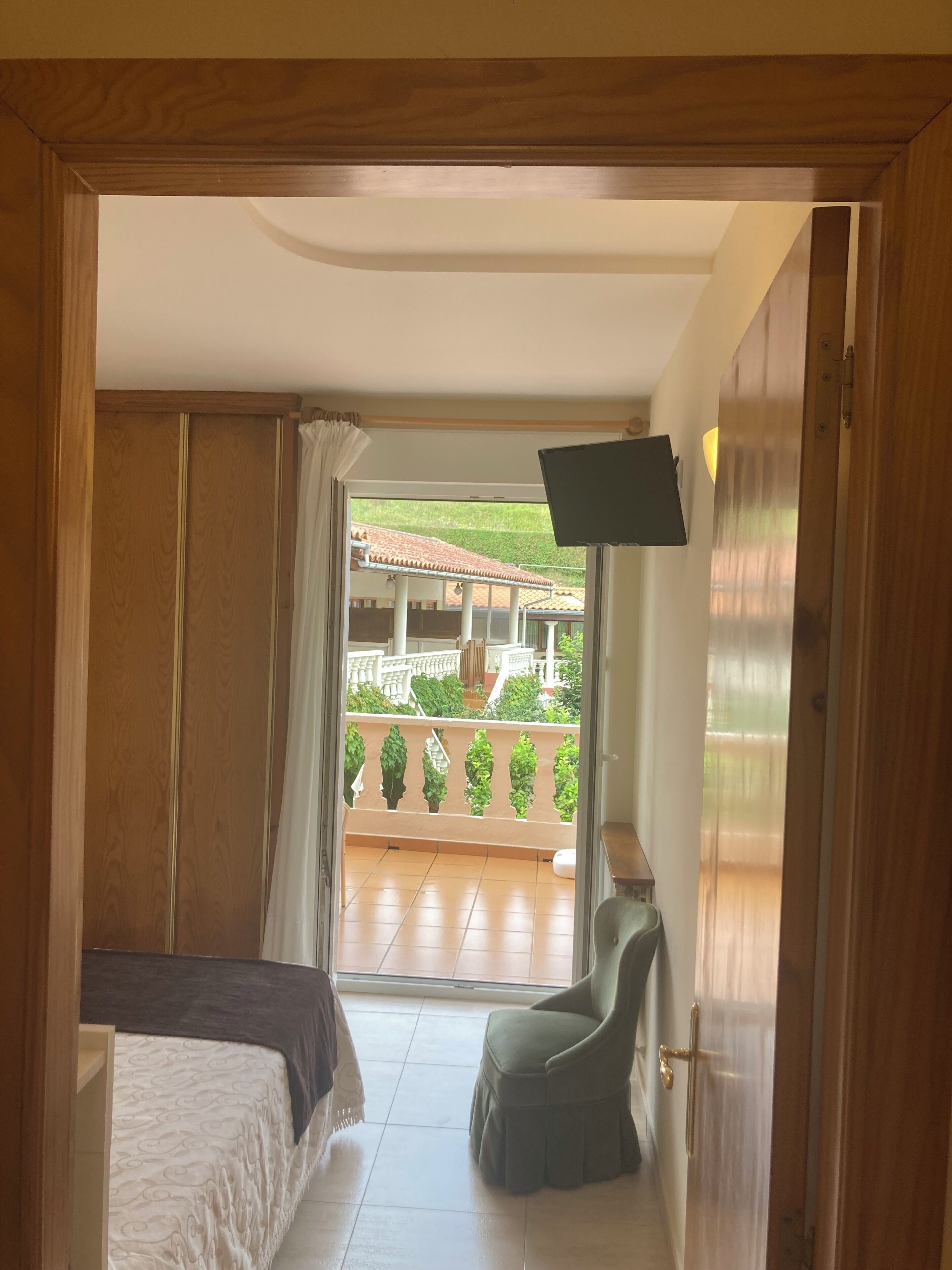 Un dormitorio con cama, una silla, un televisor y un balcón con vista a zonas verdes y un edificio.