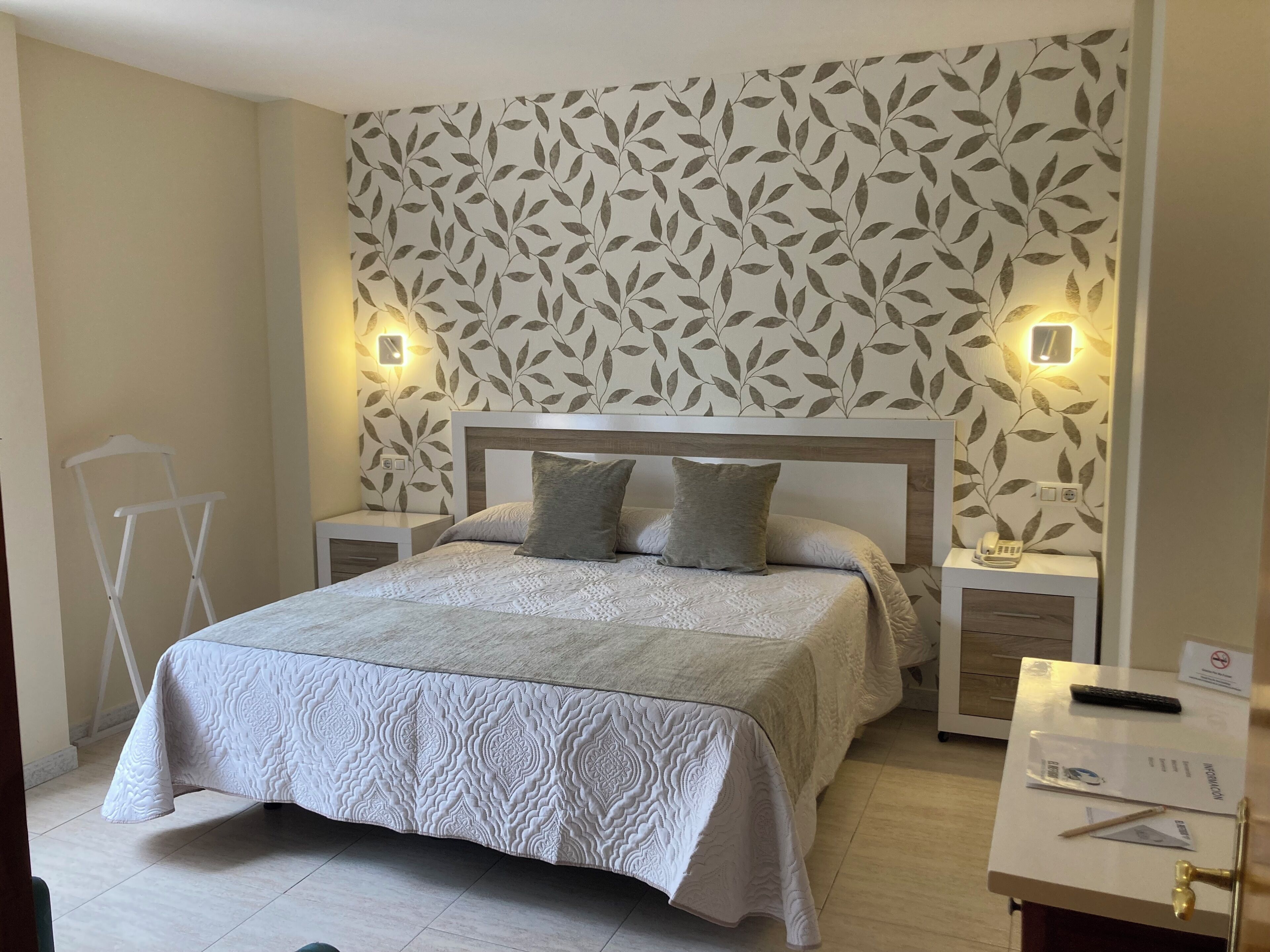 Un dormitorio ordenado con papel tapiz estampado, una cama con edredón texturizado, lámparas de noche y una mesita con teléfono y control remoto.