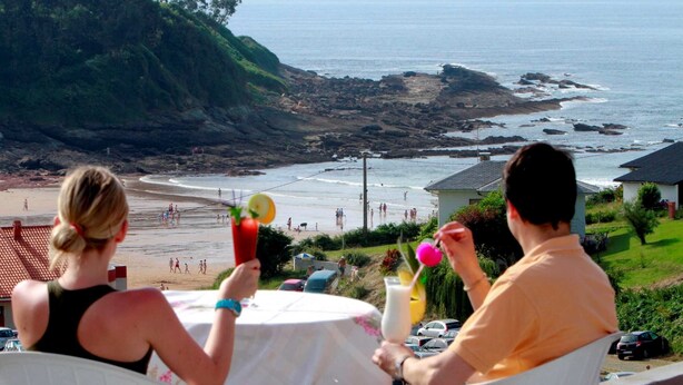 A beach view with rocky coastline, a table with drinks, and a person holding a drink. - Hotel El Refugio (Alfoz de Lloredo)