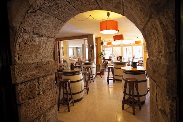 A stone archway leading to a bar area with wooden stools and barrels, warm lighting, and a view into the adjacent dining area.