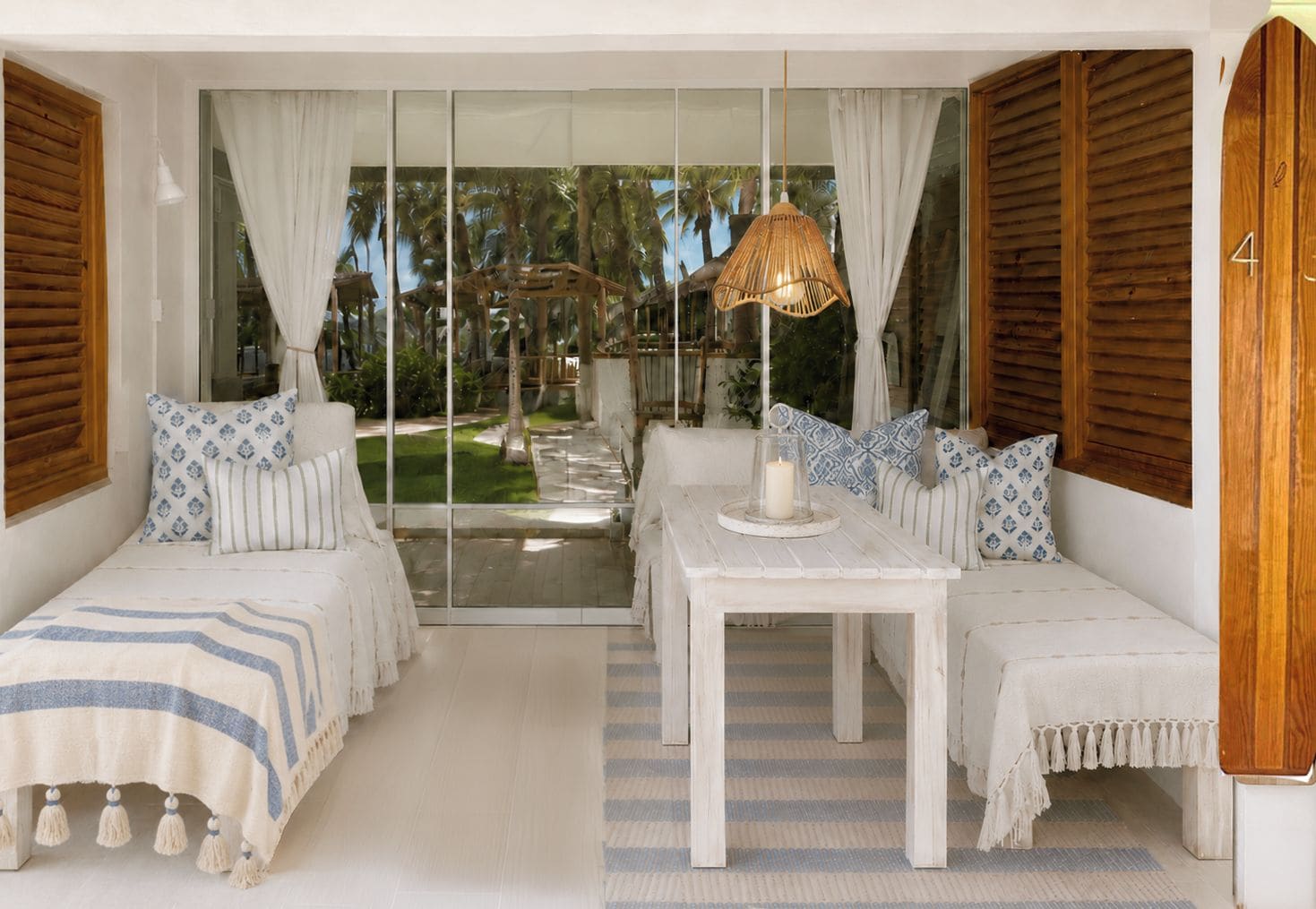A room with a glass door leading to a garden, two white sofas with patterned cushions, a small table with a candle, and a hanging rattan lamp.