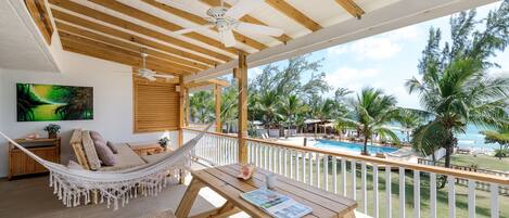 A wooden deck with a hammock, a wooden table with a magazine, and a view of a pool and palm trees.