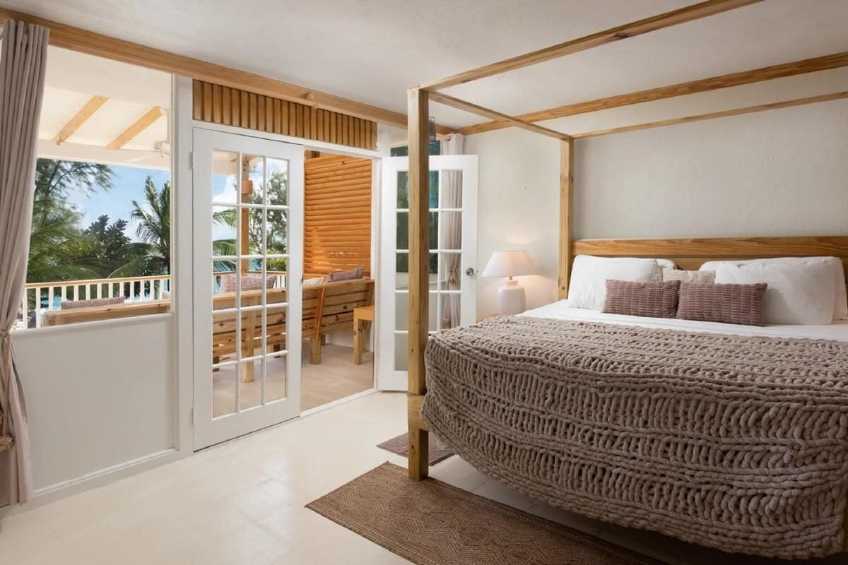 A bedroom with a large bed, a wooden headboard, a balcony with a wooden deck, and a sliding glass door leading to an outdoor area.