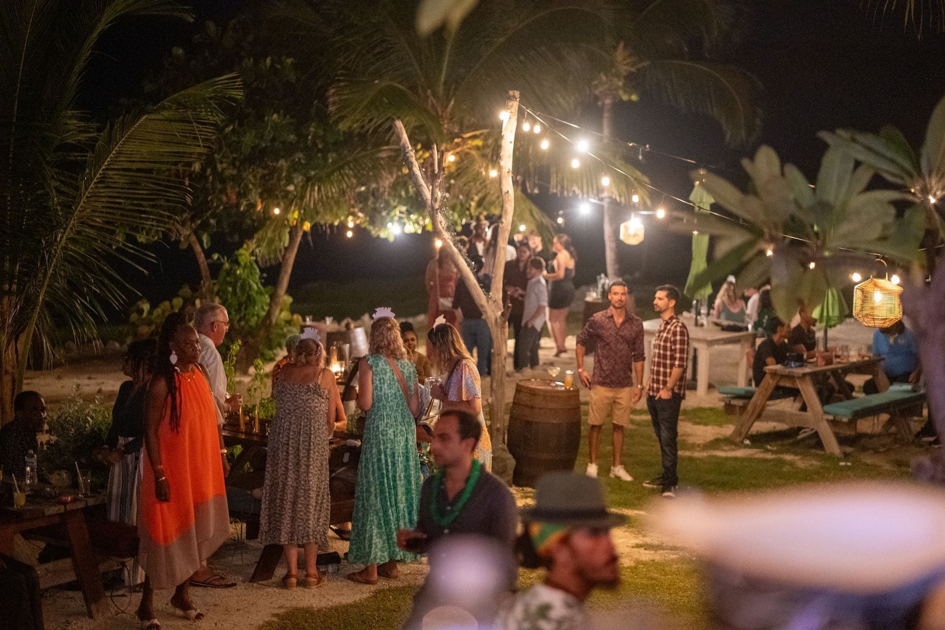 An outdoor gathering with people standing and conversing, surrounded by palm trees and string lights.