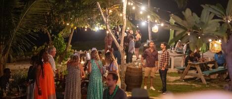 An outdoor gathering with people standing and conversing, surrounded by palm trees and string lights.
