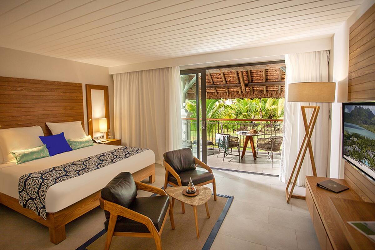 A hotel room with a large bed, two chairs, a small table, a TV, and a balcony with outdoor furniture.