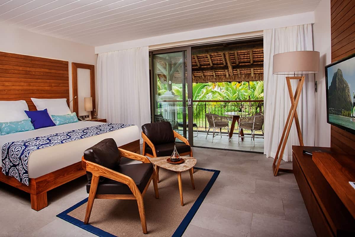 A hotel room with a bed, two chairs, a small table, a TV, and a balcony with a view of greenery.