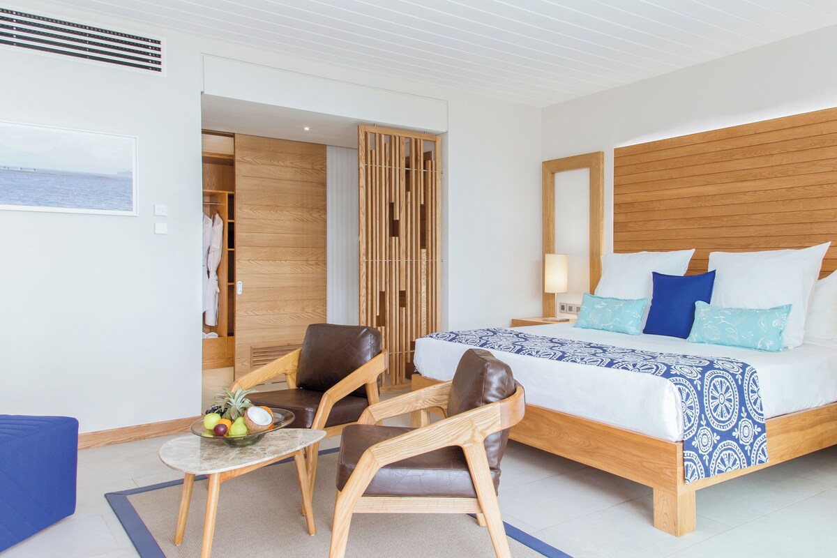 Habitación de hotel con cama, dos sillas, una mesita con frutas y vista al mar a través de una ventana.