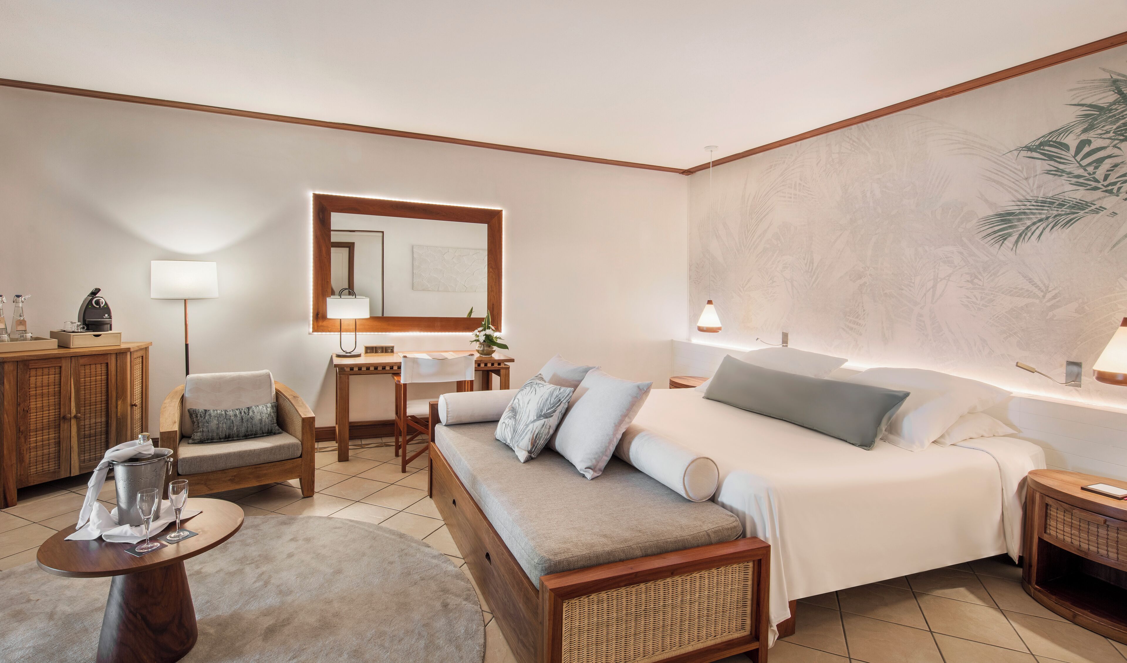 Habitación de hotel bien iluminada con una cama grande, un sofá, un escritorio y una silla. Hay un espejo grande sobre el escritorio y una pared con un diseño de planta tropical.