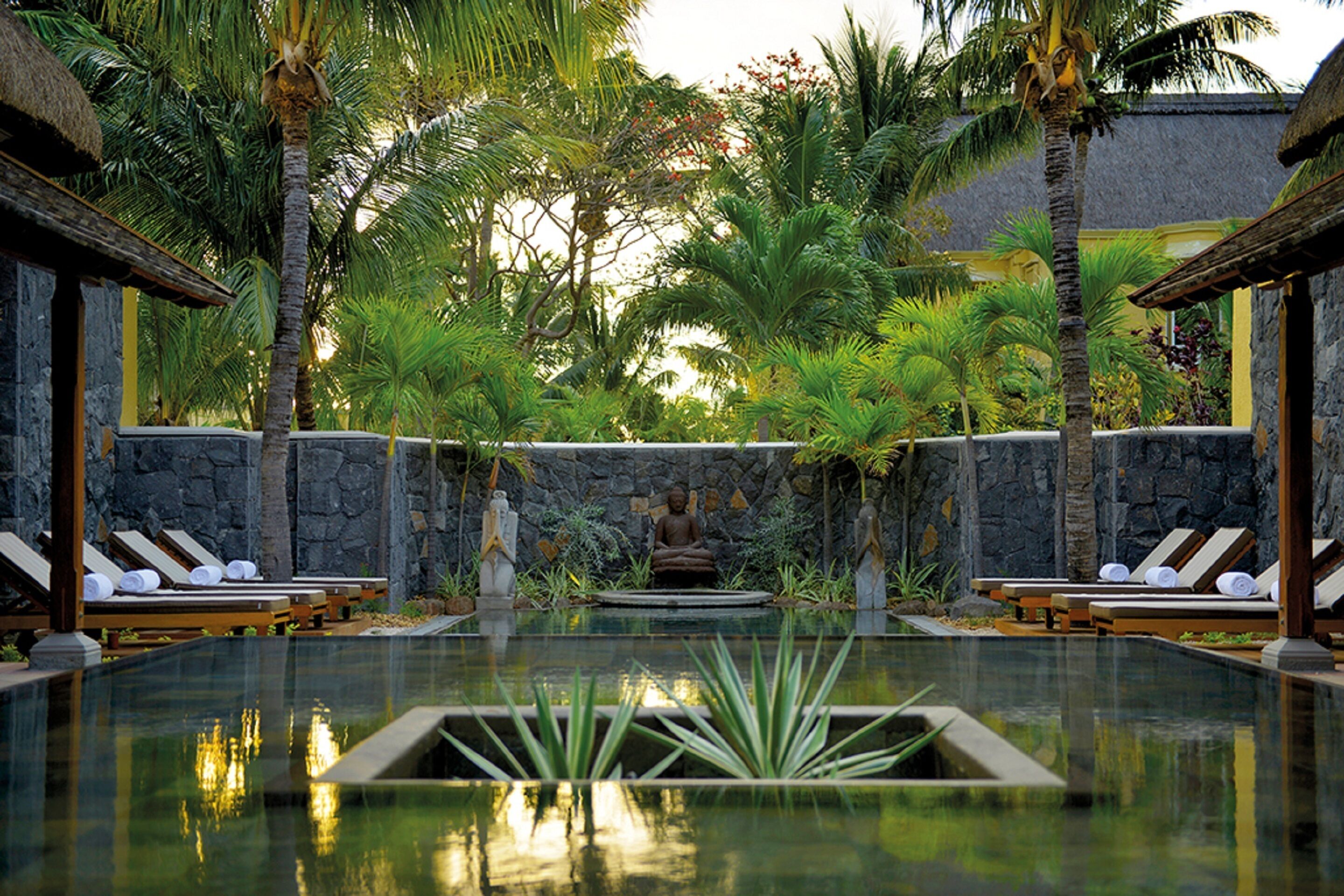 A serene pool area with lounge chairs and a Buddha statue, surrounded by tropical plants and stone walls.