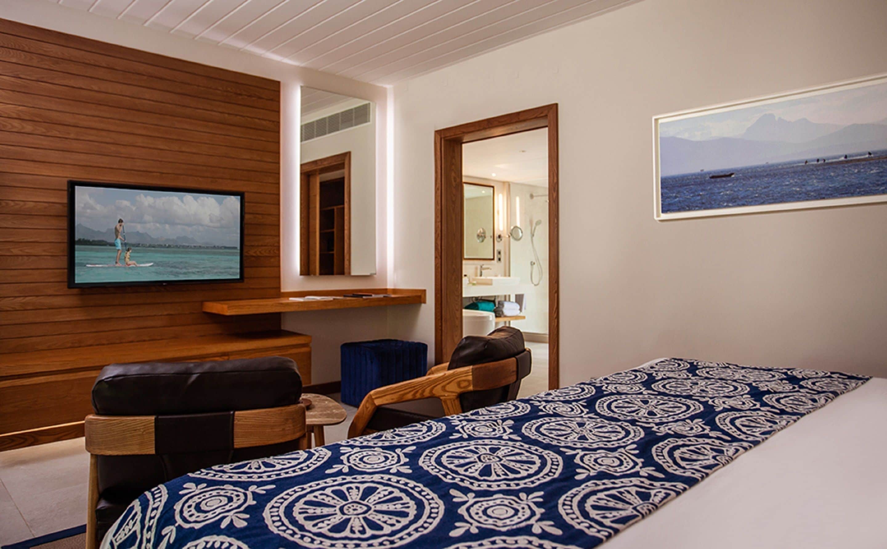 Habitación de hotel con cama, televisor montado en un panel de madera, baño con espejo y vista al mar.
