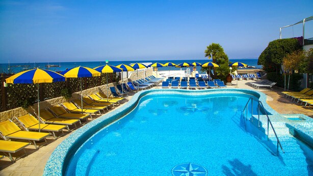 Ein Poolbereich mit blauen und gelben Sonnenschirmen, Liegestühlen und Blick auf das Meer.