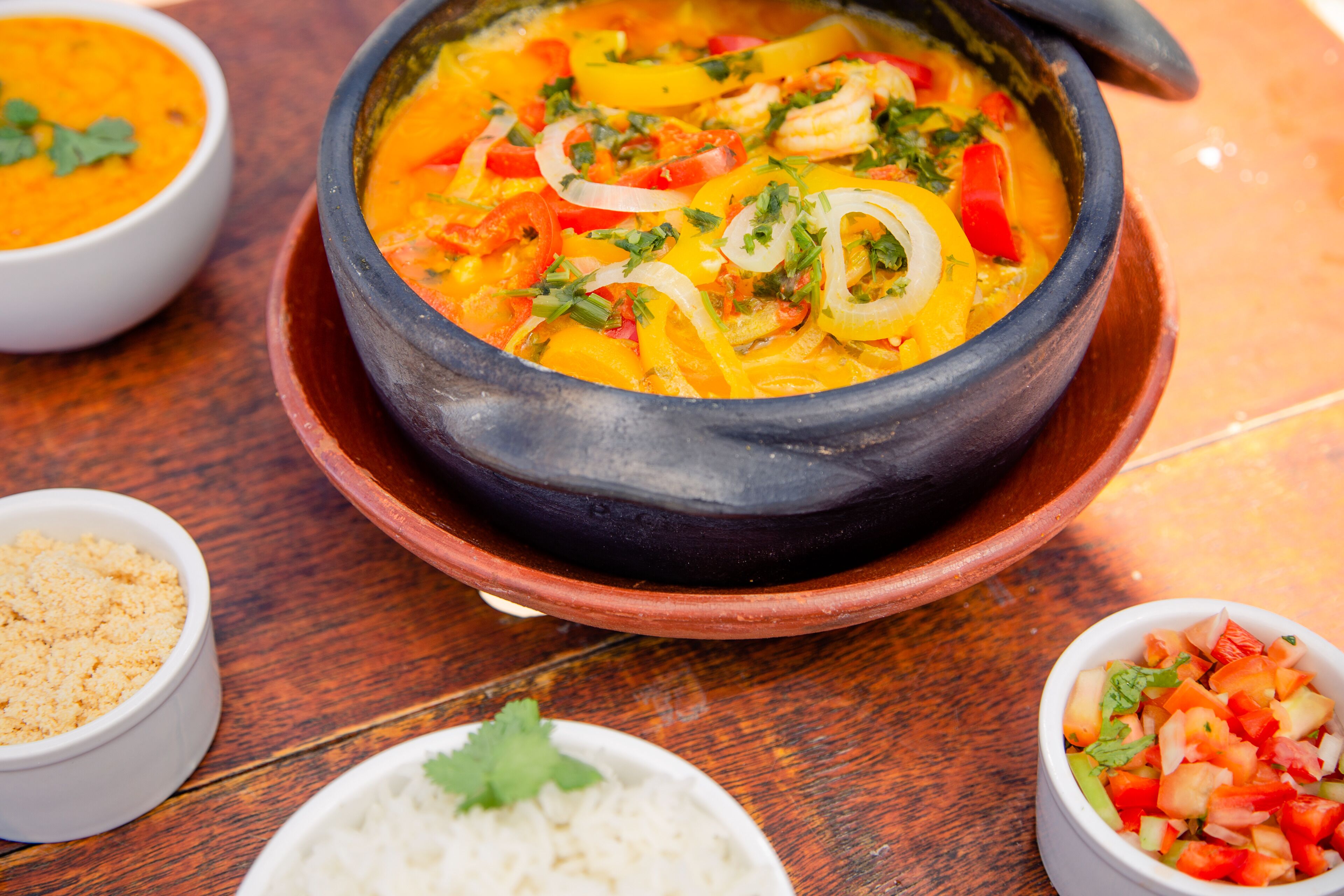 Una olla de barro tradicional llena de una sopa anaranjada vibrante, acompañada de arroz blanco y un pequeño tazón de tomates picados.