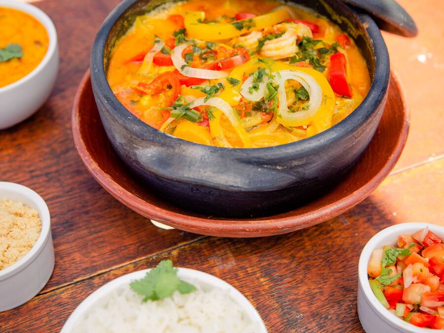 Una olla de barro tradicional llena de una sopa anaranjada vibrante, acompañada de arroz blanco y un pequeño tazón de tomates picados.