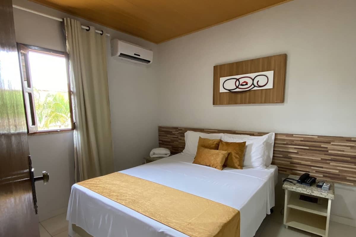 A hotel room with a bed, a nightstand, an air conditioning unit, a window with curtains, and a wall-mounted artwork.