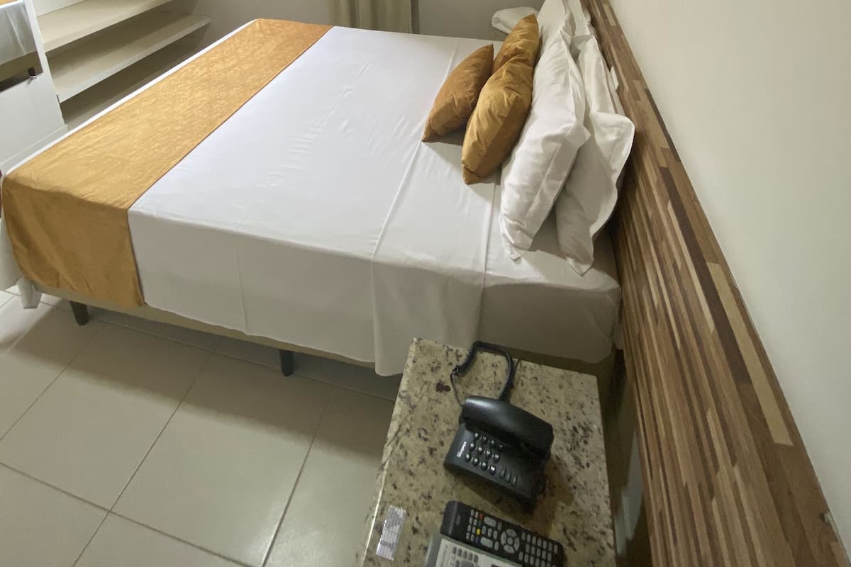 A hotel room with a bed, a wooden headboard, a window with curtains, an air conditioning unit, and a wall-mounted artwork.