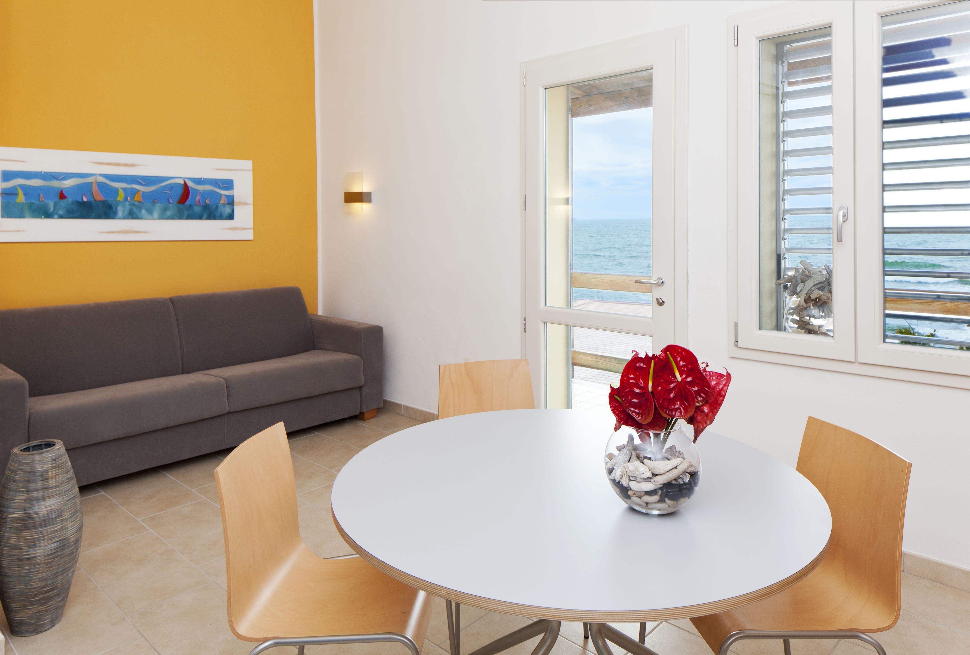Une pièce avec une table ronde, quatre chaises, un canapé gris, un vase avec des fleurs rouges, un tableau au mur et une vue sur la mer par la fenêtre.