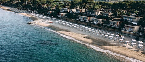 An aerial view of a coastal hotel with beachside rooms, a row of sun loungers, and a boat on the shore.