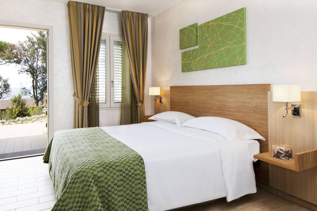 Una habitación de hotel con una cama, dos lámparas de noche, un cabecero de madera, un cuadro abstracto verde y vistas al exterior.