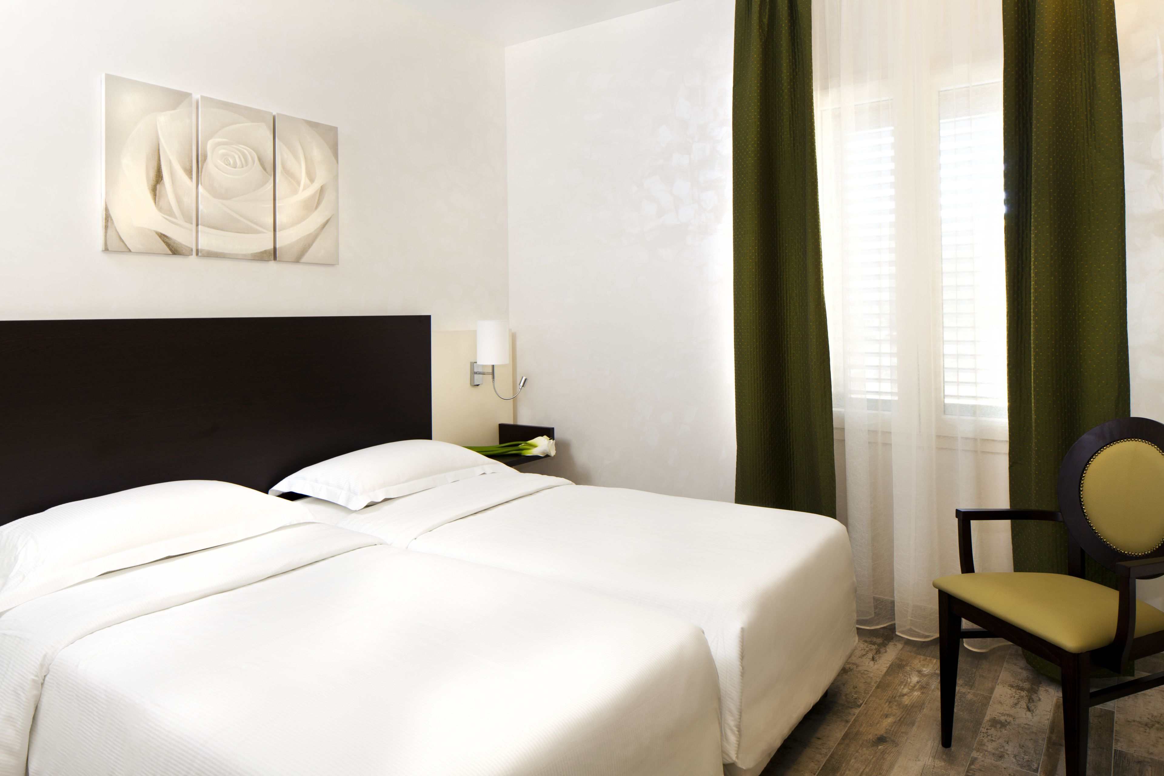 A hotel room with two beds, a nightstand, a lamp, a chair, and a large window with curtains.