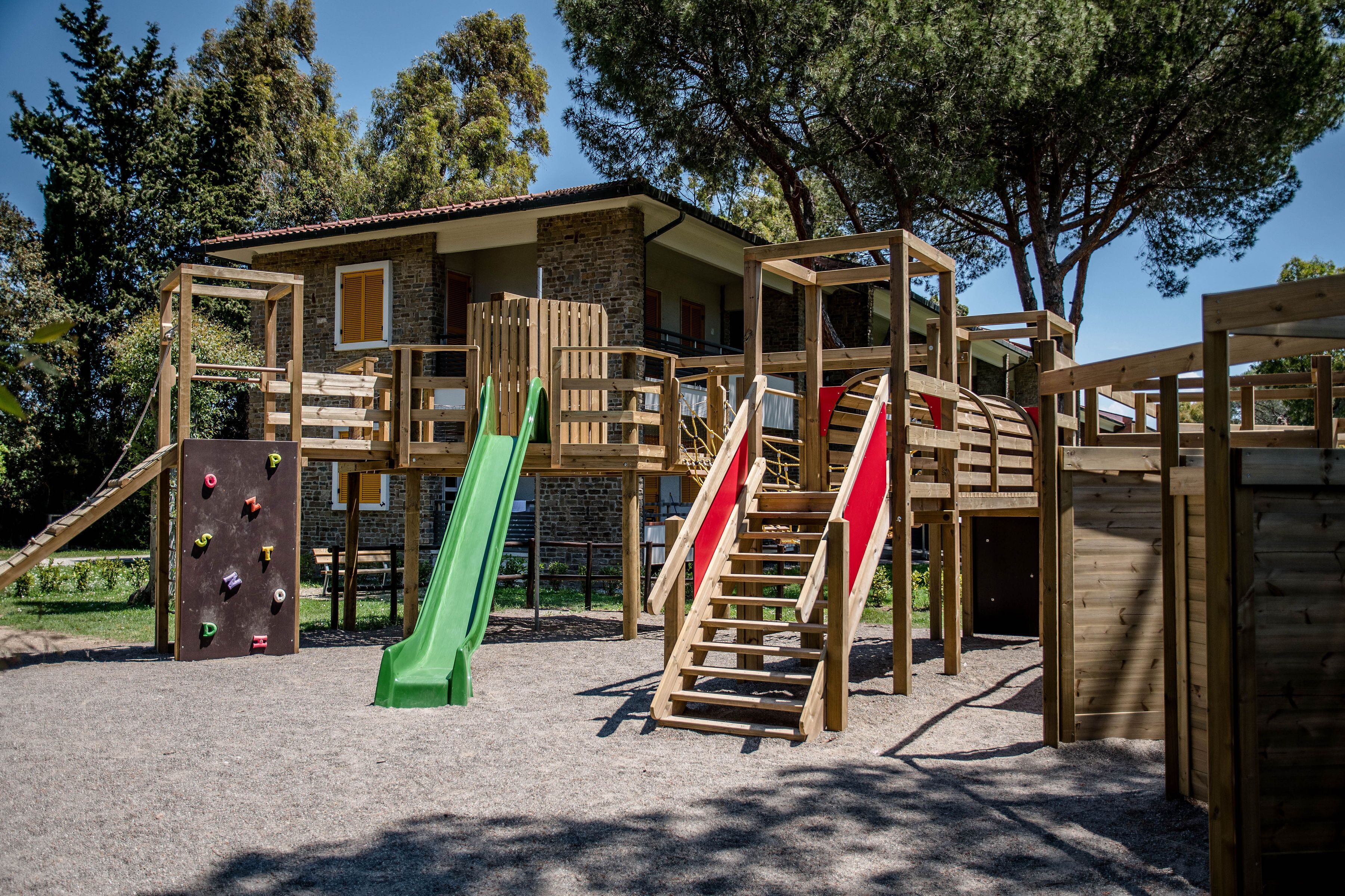 A wooden play structure with slides and climbing walls, set against a backdrop of trees and a building.