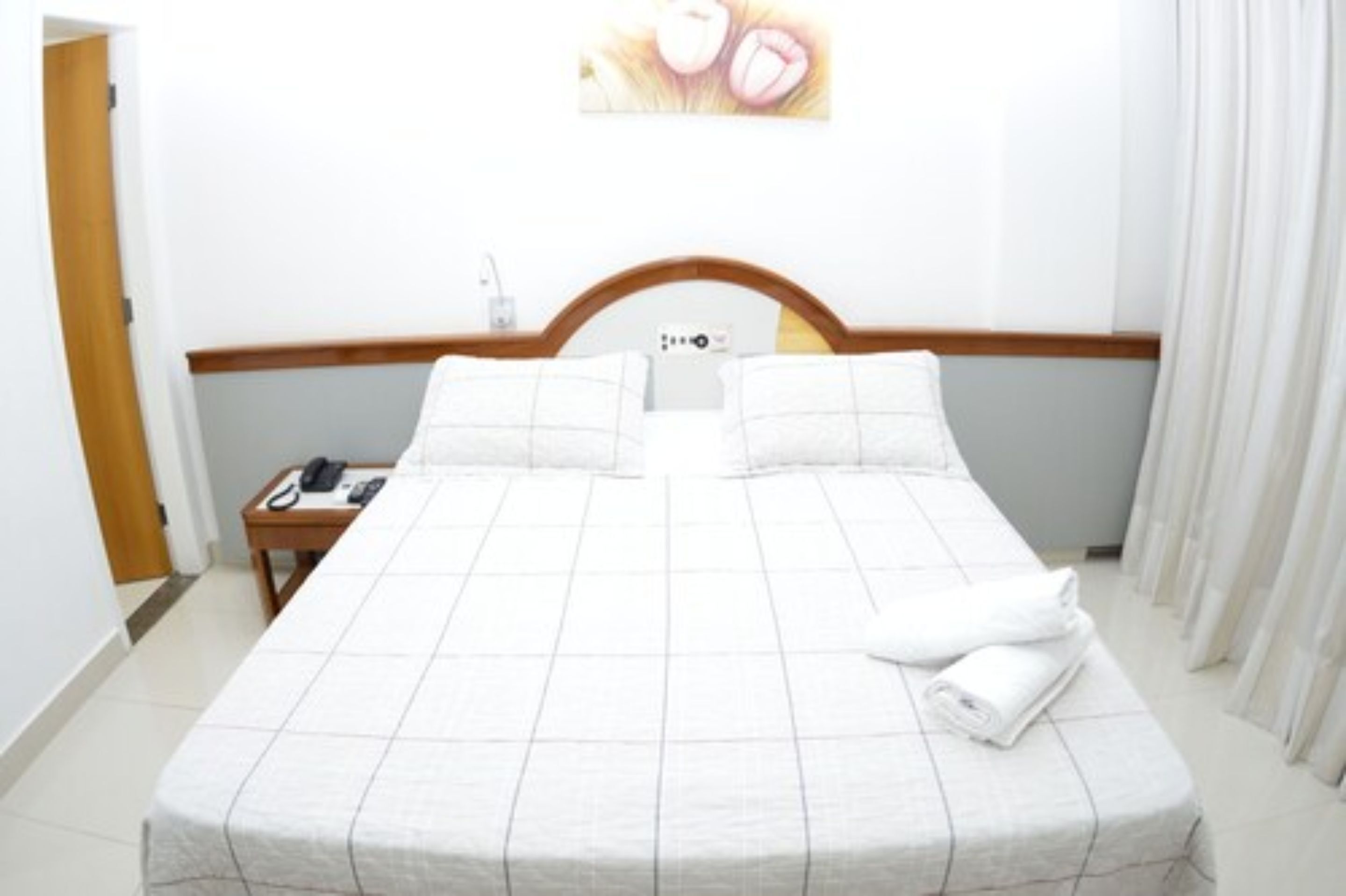 A hotel room with a bed, a bedside table with a phone, and a wall-mounted control panel.
