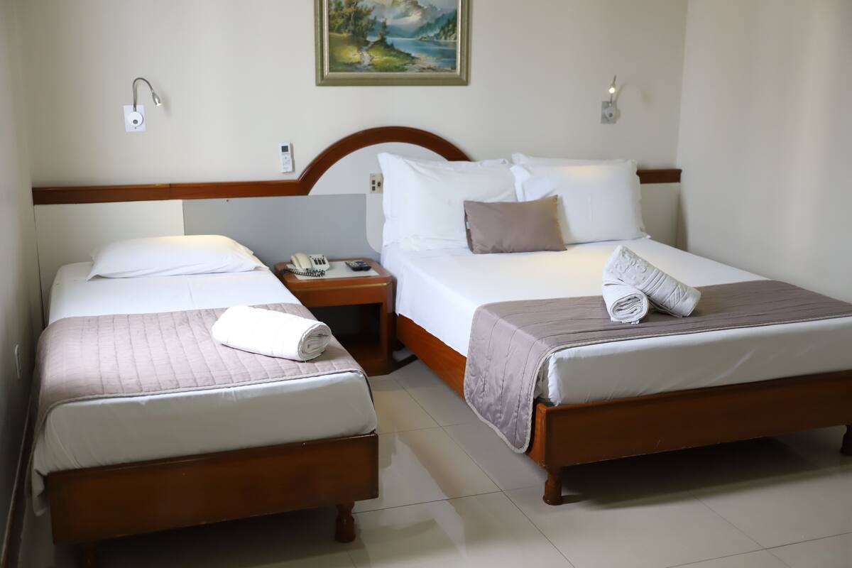 A hotel room with two single beds, a wooden headboard, a painting on the wall, and a bedside table with a phone and remote control.